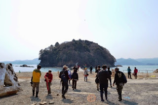 小島の見える海岸に集まり、リュックを背負って歩き出す複数の参加者たちの写真