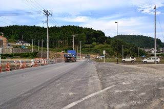 道路の左側にオレンジのバリケードが続き、青いトラックと白い車が走る舗装途中の道が山に向かって伸びている道路の写真