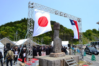 チリ国旗と日本国旗が並んで掲げられた鉄枠の下に設置されたモアイ像の後ろ姿が写っており、像の前に設置された白いテントの前にたくさんの人が集まっている写真