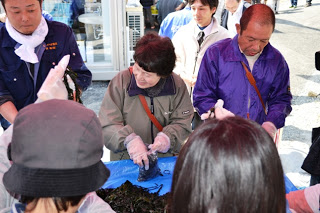 数人の人々が屋外でテーブルを囲み、手袋を着けてわかめの袋詰めに取り組んでいる様子の写真