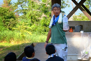 タオルを肩にかけた男性が、水が入ったペットボトルを持ちながら数人の子どもたちに説明をしている写真