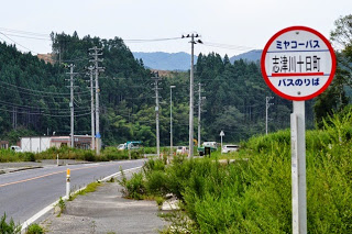 緑の森を背景に、緩やかにカーブした舗装道路沿いに「ミヤコーバス志津川十日町バスのりば」と書かれたバス停標識が立っている写真