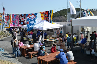 屋外のにテントやテーブル、ベンチが設置され、多くの人々が集まってにぎわっている様子を写したイベント風景の写真