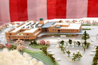 紅白幕の前に展示された、周囲に桜の木が立ち並び、駐車場が設けられた新しい病院施設の精巧な建築模型が展示されている写真