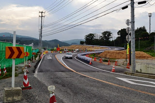 造成した高台に続く曲がりくねった道路を車が走行しており、赤色のコーンや矢印標識が多く設置されている道路工事中の写真