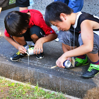 二人の男の子が空き缶に穴を開けて中の液体を溝に細く流し出している写真