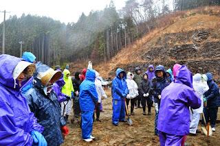 小雨が降る中、雨合羽と帽子を身につけた人々が森の中でに集まり、ところどころショベルを持参している人もいる写真