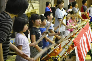 間に入った大人と沢山の子どもたちが横一列に並び、ばちを使って太鼓を叩く練習をしている様子を横から撮影した写真