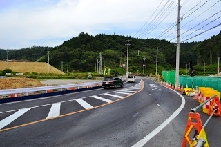 山に囲まれた緩やかなカーブの道路を2台の車が走っており、片側に緑の防音シート、もう片側に工事用資材が置かれている工事現場近くの道路の写真
