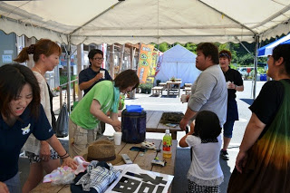 麦茶ポットやペットボトル、帽子などが置かれているにぎやかなテント下の休憩スペースで、ダンスを踊った大人や子どもがテーブルを囲みながら談笑している写真