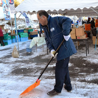 男性が雪の積もった会場の地面をオレンジ色のスコップで丁寧に除雪している、朝の準備の様子を写した写真