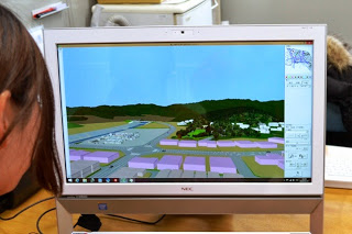 NEC製のモニターに建物や道路、緑地を含む風景の3Dモデルが表示されており、左側には画面を見ている女性の横顔の一部が写っている様子の写真