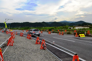片側通行の道路沿いに多くのオレンジ色の工事用バリケードが並んだ道路を車が通行している写真