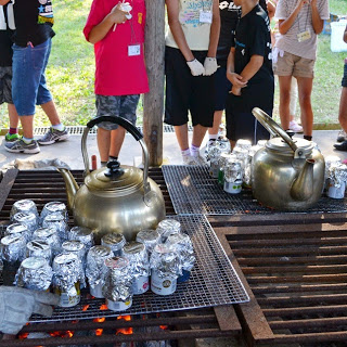 金色のやかん2つと、アルミホイルで包まれた空き缶が炭火の上の金網に沢山並べられ、数人の子どもがその様子を見守っている屋外調理の写真