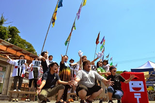 大漁旗がたなびく商店街の前で、赤色のポストくんやスタッフたちが、手を大きく広げて元気にポーズを決めている写真