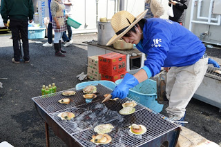 屋外で麦わら帽子をかぶった男性が網の上で殻付きのホタテを焼いている様子を写した写真