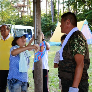 周囲には他の人たちやテントがあるキャンプ場で、青い帽子をかぶった女の子が扇子で迷彩服を着た男性に風を送ってあおいでいる写真