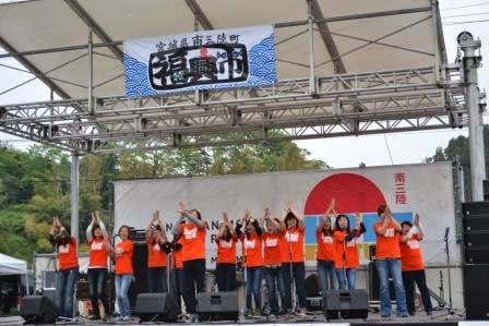 福興市と書かれたステージの上で、お揃いのオレンジ色のTシャツを着たグループが手を上げてパフォーマンスをしている写真