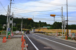 山側に向かって続く道路に車が進んでおり、両側にオレンジ色のバリケードが設置されている工事中の信号交差点の写真