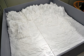 白を基調とした層状の素材で作られた山岳地形の詳細な立体模型が写っており、等高線によって標高の変化が示され、奥に向かって高い山や谷が立体的に表現された地形模型の写真