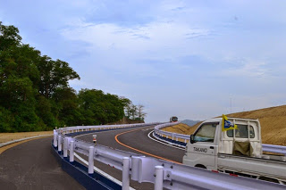 左手に木々、右手に盛土が見える、白いガードレールが両側に設置された緩やかなカーブの舗装道路を小型トラックが走行している写真