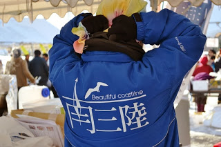 「南三陸 Beautiful coastline」と書かれたジャンパーを着た人がテントの下で手ぬぐいを頭に巻いている後ろ姿を写した写真