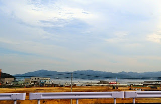 ガードレール越しの復興工事中の町とその先に広がる海が見え、遠くに山並みが続く風景の写真