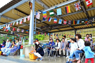 国旗が飾られた屋根の下で、観客たちが椅子に座ったり立ち上がったりして競技を見守っている写真