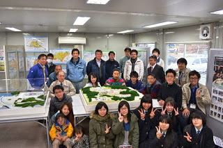 壁には地図やポスターが掲示されている室内で、複数の人物が模型やジオラマが置かれた展示テーブルの周りに集まっている写真