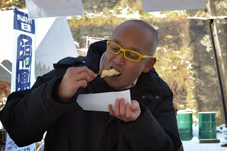 黄色いメガネの男性が串に刺さった揚げ物をほおばっている写真