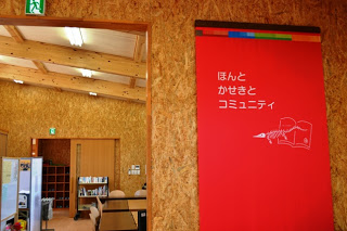 壁に赤い縦長の看板がかかっていて「ほんとかせきとコミュニティ」と白字で書かれており、開放的な室内空間となっている入り口前を写した写真