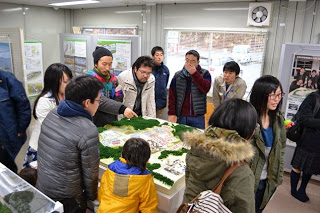 壁にはポスターや情報展示、詳細な模型やジオラマが置かれた展示室内で、冬服を着た複数の人達が模型を囲んで立っている様子が写っている写