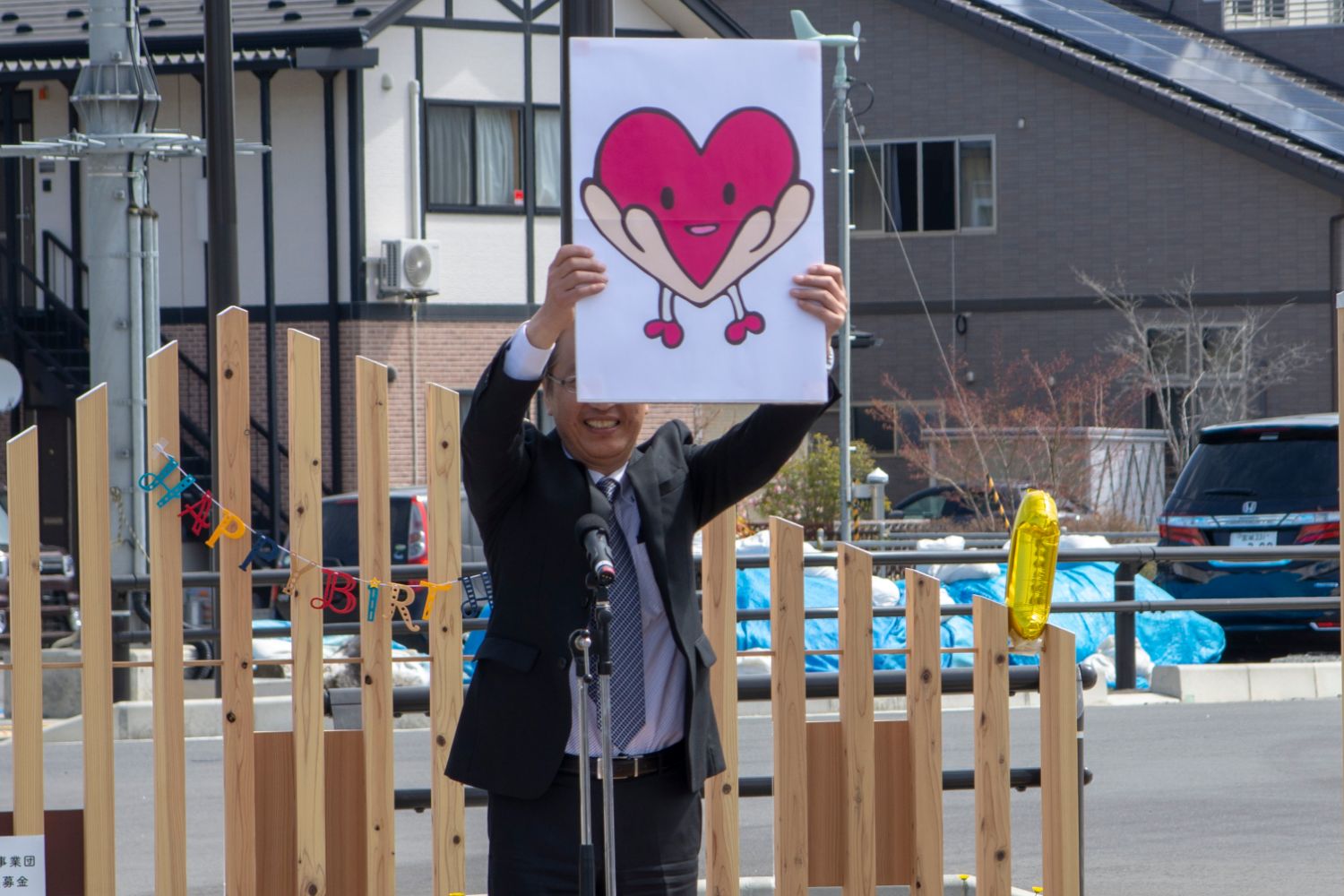 スーツ姿の男性がマイクの前でハート型のキャラクターが描かれた紙を笑顔で高く掲げている発表の様子の写真