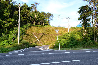 幹線道路から分かれ、草に覆われた法面が見える山の斜面に沿って曲がる緩やかな坂道の写真