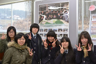 大きなポスターの様な物が飾られた室内に、手でピースサインをしている6人の女子生徒が立っている様子が写っている写真