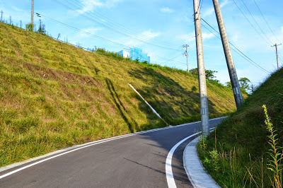 草に覆われた斜面に沿って、右に向かってカーブした舗装された緩やかな坂道が続いている様子の写真