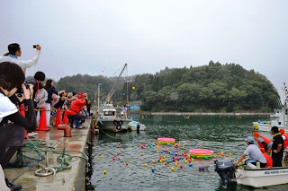 港の海上にカラフルなボールと浮き輪が浮かび、岸からたくさんの人が眺めたり写真を撮ったりしている様子の写真