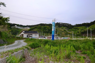 遠くに低い山々が見える緑豊かな草地の中に青い柱状の案内表示が立っている様子を写した写真