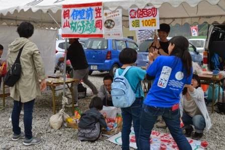 屋台の下で子どもたちがゲームに参加し、周りの大人たちが見守ったり写真を撮ったりしている様子の写真