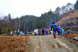 分厚い雲が広がる空の下、色とりどりの雨具を着た人々が、霧がかった山の森の中に集まっている様子の写真