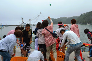 奥に漁船や湾が見える港の岸壁で、たくさんの人がカゴに向かってカラーボールを投げ入れている様子の写真