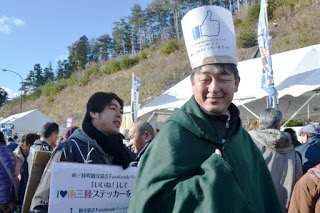 屋外のイベント会場でFacebookの「いいね！」マークが大きく描かれた白い紙製の帽子をかぶった山平さんが緑色のコートを着て立っており、その後ろにボードを首にかけた男性が立っている写真