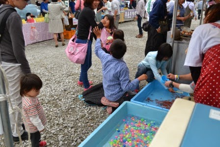 周囲に家族連れが集まり、屋台で水色の水槽に浮かぶ色とりどりのボールをすくうゲームをしている子どもたちの様子の写真