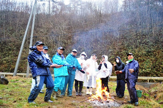 葉っぱのない木々が立つ斜面と、ガードレールが設置された道路を背景に、雨具を着た9人の参加者が、屋外で焚き火を囲んでいる写真