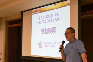 「吸う人も吸わない人も一緒に考えてみませんか？受動喫煙」と書かれたスライドを背景に、マイクを持った男性講師が話している写真