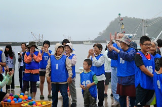 青いゼッケンを着た子どもから高齢者までの参加者たちが岸壁で手を叩いたり歓声を上げている様子の写真