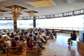 海が見える明るいラウンジで、多くの参加者がが椅子に座り、前方でマイクを持った男性の話を静かに聞いている写真