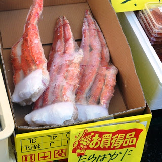 大きな冷凍のタラバガニの足が段ボール箱に詰められ、「お買得品」と表示された黄色い値札とともに並べられている写真