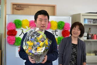 入所式と書かれたホワイトボードの前で、女性が見つめるなか、透明のビニールがかぶせられた黄色や青色の花のアレンジメントフラワーを両手で持ち挨拶をしている男性の写真