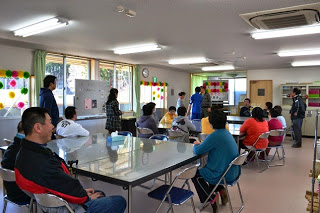 窓ガラスから太陽の光が差し込んだ明るい室内で、テーブルを囲んで置かれた椅子に人々が座り、前方に座っている男性の話を聞いている様子の写真